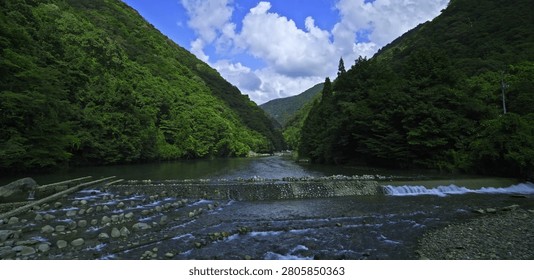 This winding river flows through a vibrant green valley between dense forest mountain hills under a bright blue sky filled with white clouds in this tranquil natural landscape scene