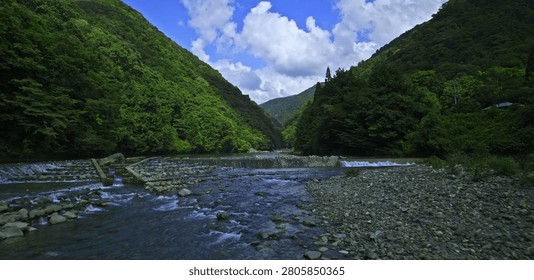 This winding river flows through a vibrant green valley between dense forest mountain hills under a bright blue sky filled with white clouds in this tranquil natural landscape scene