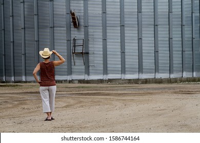 This Woman Farmer Is Standing With Her Hand On Her Cowboy Hat Looking At A Metal Grain Bin.