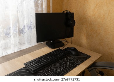 This workspace has a black computer monitor, keyboard, mouse, and headphones placed neatly on a desk in a well-lit room. Daylight comes through a window with white curtains.
