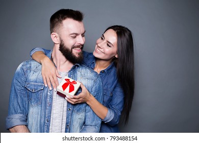 This Is For You My Dear Husband. A Beautiful Happy Woman Presents A Gift To Her Beloved And Hugs Him From Behind On A Gray Background. Valentine's Day.