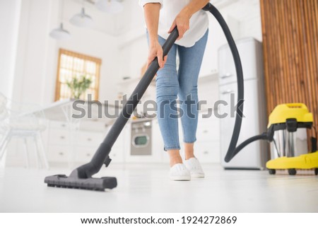 Thorough cleaning. Slender woman in jeans and moccasins doing afternoon vacuuming in white kitchen, no face