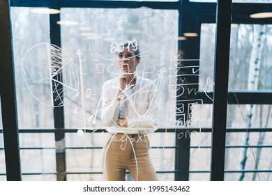 Though glass view of smart female in office clothes looking at data on transparent board while working and making decision