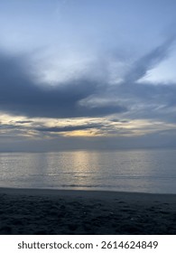 Though the sun is concealed by clouds, its light spreads across the sky in a warm, glowing hue. The calm ocean reflects the dusk tones, creating a scene of soft beauty and peace.

