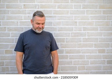 Thoughful Man Looking Down To The Ground With A Smile As He Stands In Front Of A White Brick Wall