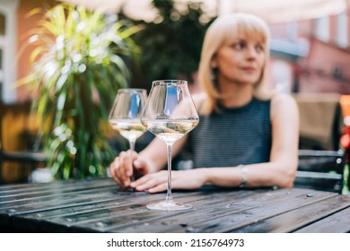 Thoughtful Adult Mature Woman Sitting In Bar Outdoors With Wine Glasses And Blurry Restaurant Background Scene, Drinking White Wine. Summer Sunny Day On Patio. People Lifestyle