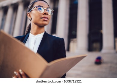 Thoughtful African American businesswoman in optical spectacles holding documents looking away while standing in urban setting with copy space. Female dark skinned student of high economic university