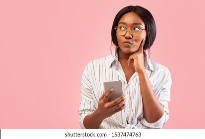 Thoughtful African American Girl Using Smartphone Thinking About Something Standing Over Pink Studio Background. Free Space