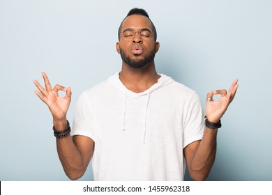 Thoughtful African American man in glasses calming, breathing deep, meditating with closed eyes, mudra gesture, young male practicing yoga, doing exercise, standing isolated on studio background