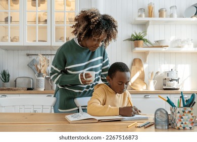 Thoughtful African American mother of pupil boy approaches studying pensive son with mug of tea check completion of school assignment. Caring black mom support son in difficult education task at home - Powered by Shutterstock