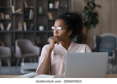 Thoughtful African American woman in glasses looking to aside touching chin, pondering project strategy or ideas, pensive young female student or freelancer sitting at work desk with laptop