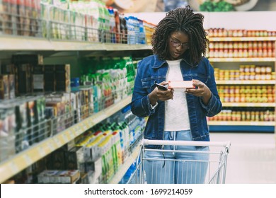 Thoughtful African American Woman Shopping At Grocery Store. Serious Young Customer Standing With Shopping Cart And Reading Information On Packaging. Shopping Concept