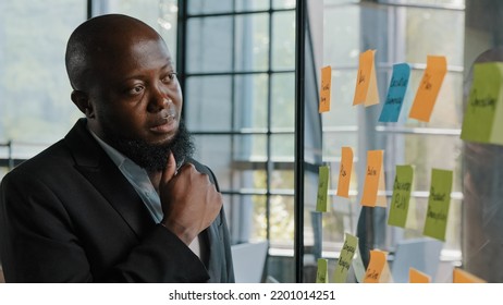 Thoughtful African Entrepreneur American Adult Bearded Male Businessman Team Leader Stand Alone Look At Transparent Glassy Wall With Paper Sticky Notes Managing Goals Project Planning Effective Work