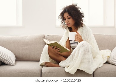 Thoughtful african-american student girl reading book. Young woman studying at home, sitting on beige couch wrapped up in white blanket and holding coffee cup, copy space - Powered by Shutterstock