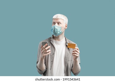 Thoughtful albino guy in face mask using credit card and smartphone to shop online on turquoise background. Young man buying goods on internet during coronavirus lockdown - Powered by Shutterstock