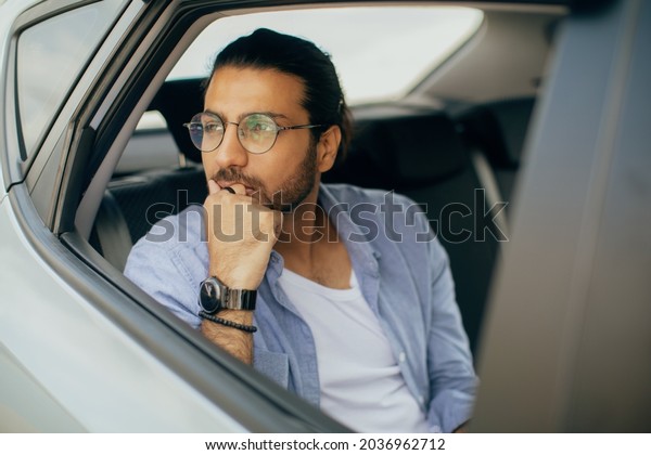 Thoughtful arab guy sitting in car, dreaming about\
something while going somewhere by taxi, pensive middle-eastern guy\
passenger looking through window, leaning on his hand, copy\
space