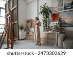 Thoughtful artist woman with inspiration paints picture, pondering ideas, holding in hands palette with oil paints in front of easel with canvas. Focused female standing barefoot in cozy art studio. 