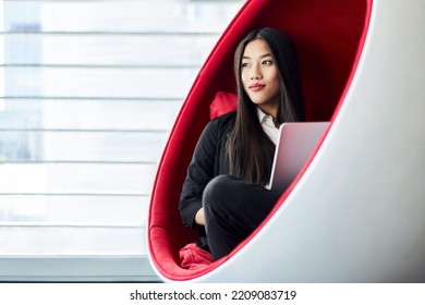 Thoughtful Asian Business Woman Sitting Inside Ball Chair At Office Using Laptop