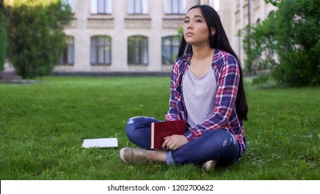 Thoughtful asian female student sitting on lawn alone and missing parents