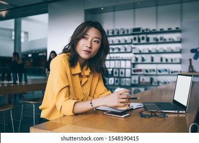 Thoughtful Asian Female Worker In Yellow Blouse At Desk With Laptop On At Coffee Break In Electronics Store In Hong Kong City