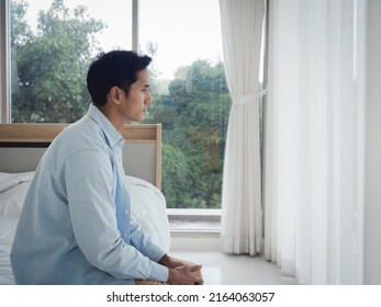 Thoughtful Asian man in denim shirt sitting on the bed feeling lonely, looking away with sadness and thinking in the bedroom at home near the glass window in the morning.