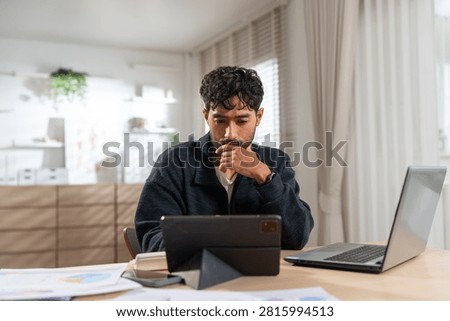Thoughtful Asian man using tablet computer analyzing financial data at home, serious male freelancer looking tablet near laptop sitting desk remote office