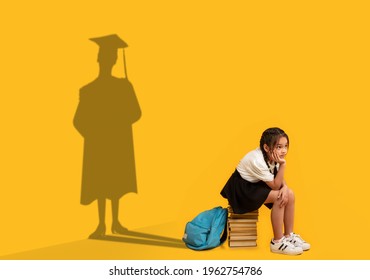 Thoughtful Asian Schoolgirl Sitting On Stack Of Books, Dreaming Of College Graduation, Shadow Of University Student In Mortarboard And Mantle On Orange Studio Wall. Success And Potential Concept