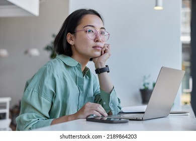 Thoughtful Asian Woman Works Freelancer Uses Laptop Computer Smartphone Has Distance Job Poses Against Cozy Interior Indoors Wears Spectacles And Shirt. Female Digital Nomad Enjoys Modern Lifestyle