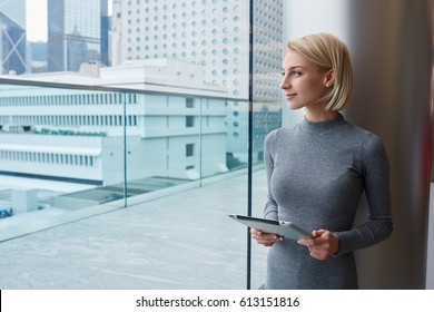 Thoughtful Attractive Blonde Female Administrative Manager Holding Modern Touchpad Standing Near Window In Coworking On Urban Background, Charming Pensive Hipster Girl Admire Cityscape From Office