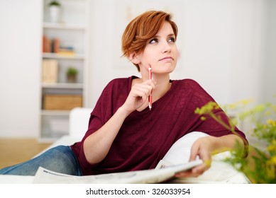 Thoughtful Attractive Young Woman Answering Crossword Puzzle Game On Newspaper At The Living Room Couch.