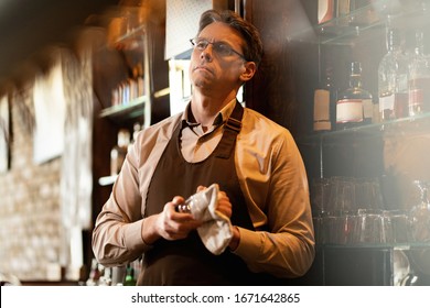 Thoughtful bartender working at bar counter and cleaning drinking glass.  - Powered by Shutterstock