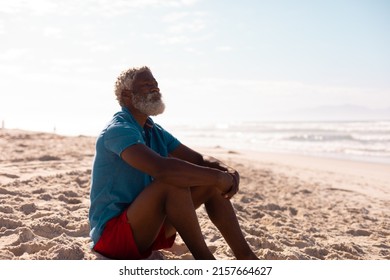 Thoughtful bearded african american senior man with gray hair sitting on sandy beach against sky.  - Powered by Shutterstock