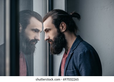 thoughtful bearded man looking at reflection in glass