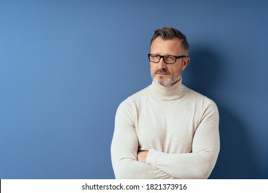 Thoughtful Bearded Middle-aged Man In Glasses And White Turtleneck Sweater Standing With His Arms Folded Against Blue Background With Copy Space And Looking Away