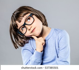 bedachte schöne kleine Mädchen mit ernsthaften Brillen, die ihren Kopf halten, um sich vorzustellen, zu denken, zu zögern oder eine intelligente Idee oder Lösung zu haben, grauer Hintergrund-Studio