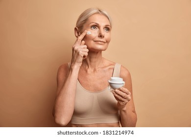 Thoughtful beautiful mature woman applies face cream has anti aging treatment wears cropped top has light hair concentrated somewhere isolated on beige background. Senior lady wants to reduce wrinkles - Powered by Shutterstock