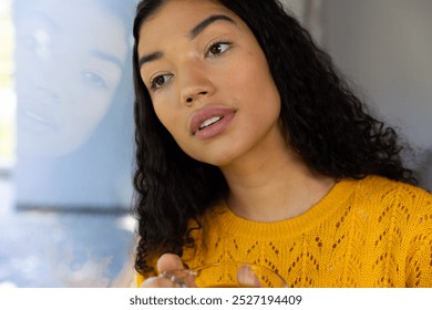 Thoughtful biracial woman holding cup of tea and looking out window at home. Lifestyle, free time and domestic life, unaltered. - Powered by Shutterstock