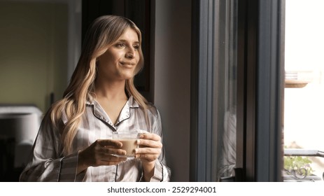 Thoughtful blond smiling transgender woman holding fresh coffee cup while looking through window at home - Powered by Shutterstock