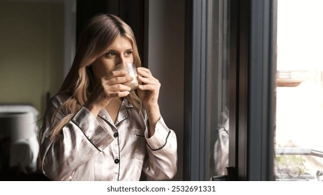 Thoughtful blond transgender woman in nightwear drinking fresh coffee as she looks through window while standing at home - Powered by Shutterstock