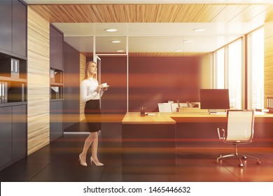 Thoughtful blonde businesswoman standing in stylish office with gray and wooden walls, tiled floor, gray and wooden computer desk and bookcases with folders. Toned image