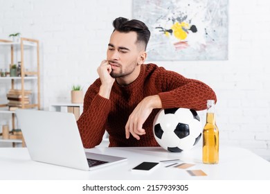 thoughtful bookmaker with soccer ball looking at laptop near smartphone and credit cards - Powered by Shutterstock