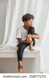 Thoughtful bored and sad indian boy sitting on a table barefoot, looking nowhere. Against white curtain. - Shutterstock ID 2189771751