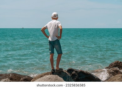 Thoughtful boy in cap, white shirt and shorts stands on the rocks by the sea