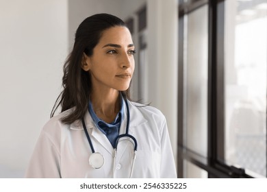 Thoughtful Brazilian female doctor in white coat with stethoscope gazing away with serious expression, thinking on medical specialist career, looking out window, deep in thoughts. Future, medicine - Powered by Shutterstock