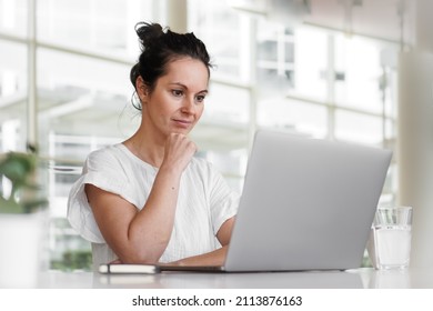 thoughtful brooding remote working dark haired woman sitting infront of a laptop or notebook in casual outfit on her work desk in her modern airy bright living room home office with many windows