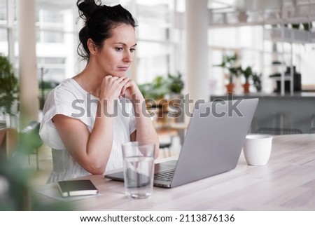 thoughtful brooding remote working dark haired woman sitting infront of a laptop or notebook in casual outfit on her work desk in her modern airy bright living room home office with many windows