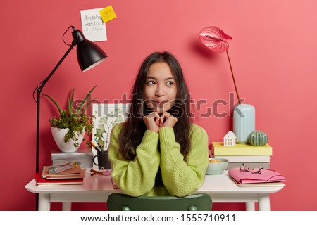 Thoughtful brunette Asian woman with hands under chin, being deep in thoughts, rests after preparing home assigment, sits at chair against working place, pink wall, creative written sticky notes