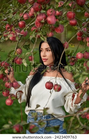 Thoughtful brunette woman hiding in tree branches, laden with red apples. Portrait of Caucasian female wearing vivid necklace, posing in apple orchard in early autumn. Harvest, female beauty concept.