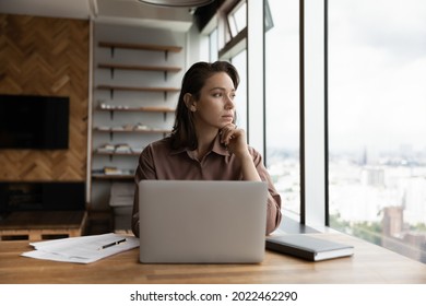 Thoughtful business owner woman looking out of window at city, sitting at workplace with laptop, thinking over solutions, challenges, company future vision, making decision, feeling uncertain