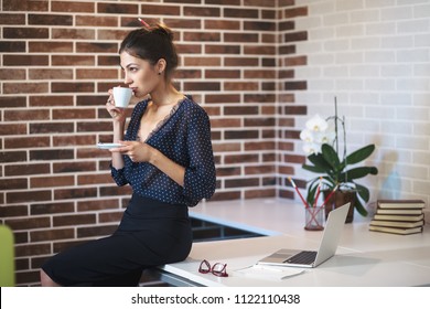 Thoughtful business woman, drinking a cup of coffee while sitting on the table in the office, wearing blouse with dots and pencil skirt looking to right, over brick wall background.